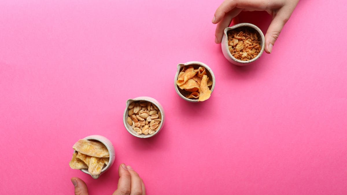 Top view of crop anonymous people taking bowls of dried banana and fruit for healthy snack on pink background